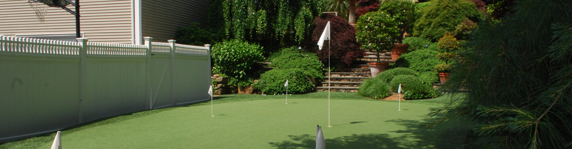 Backyard putting green with five white flag pins beside a basketball hoop, white vinyl fence, and tiered garden landscaping with Japanese maples and tropical plants
